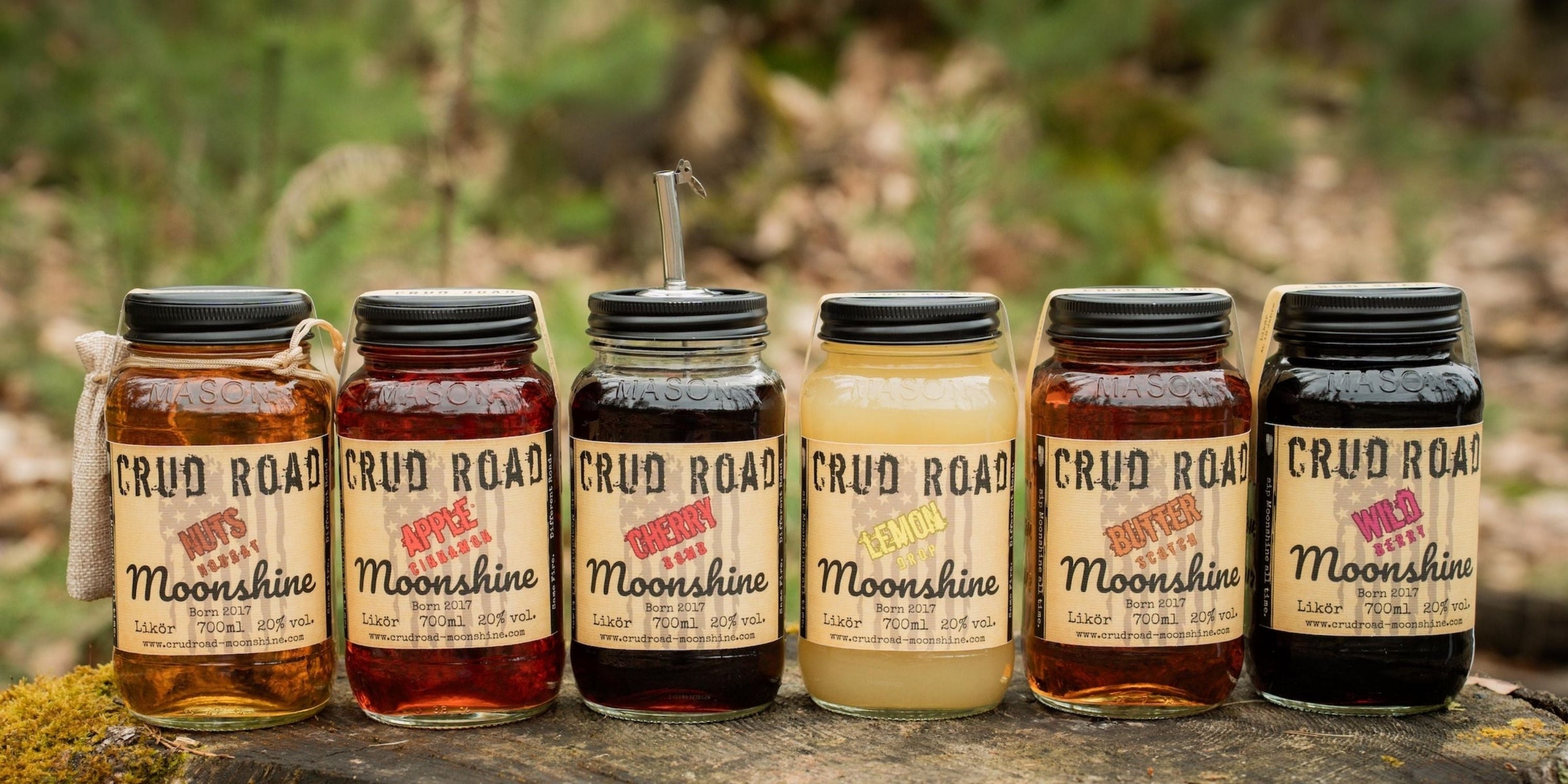 Vier Flaschen Crud Road MOONSHINE mit amerikanischer Flagge auf dem Etikett, aufgestellt auf einem Tisch, mit Angaben zu Inhalt (700ml) und Alkoholgehalt (20% vol.).
